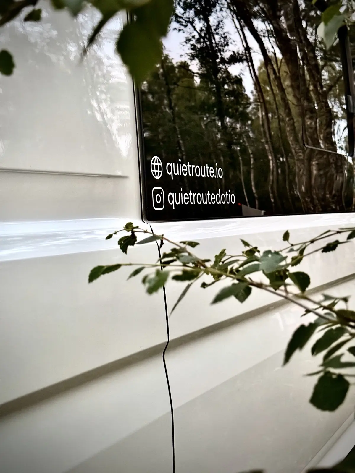 A quietroute.io sticker on the side window of the campervan, birch trees reflected in the glass