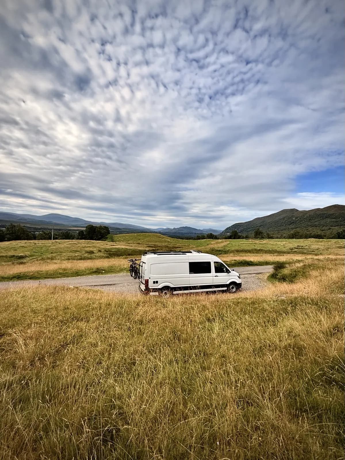 A white VW Crafter campervan with bikes on the back, parked on a track through moorland grass with hills behind