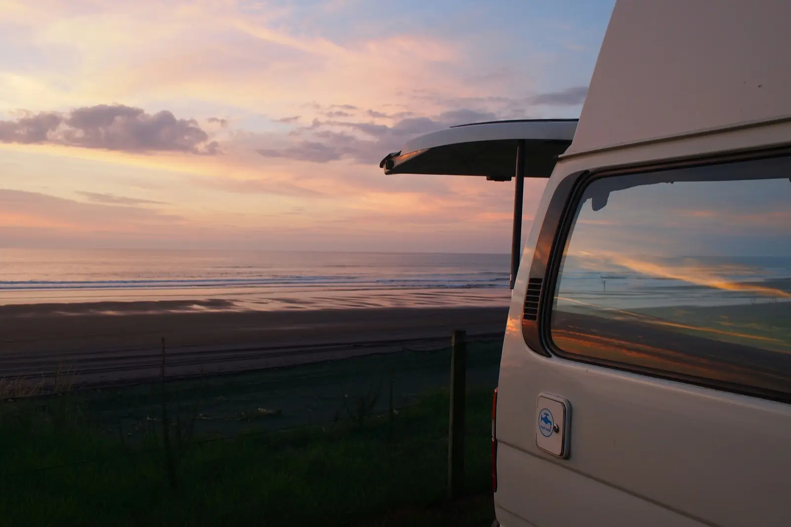 White campervan parked beside an empty beach at sunset, pink and orange sky reflecting in the side window