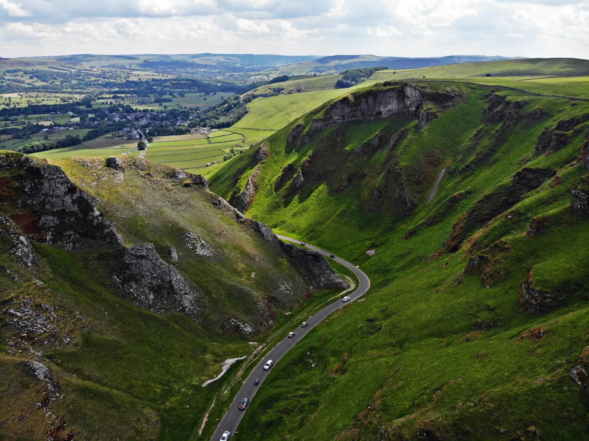 Aerial view of Winnats Pass in the Peak District, the road threading between green limestone ridges with the village of Castleton and a patchwork of fields in the valley beyond.