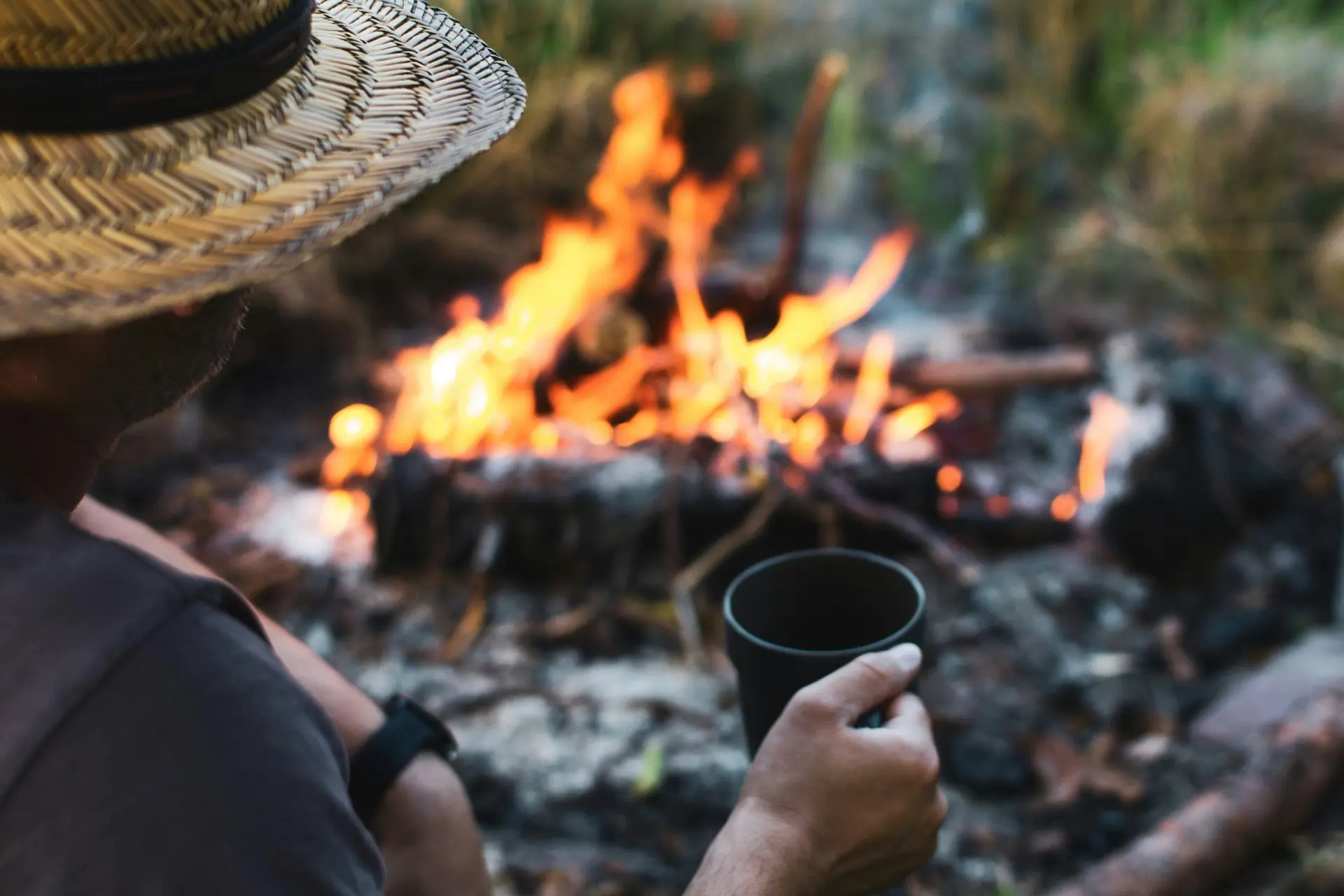 View over the shoulder of a person in a straw hat holding a dark enamel mug, small campfire burning in front of them.