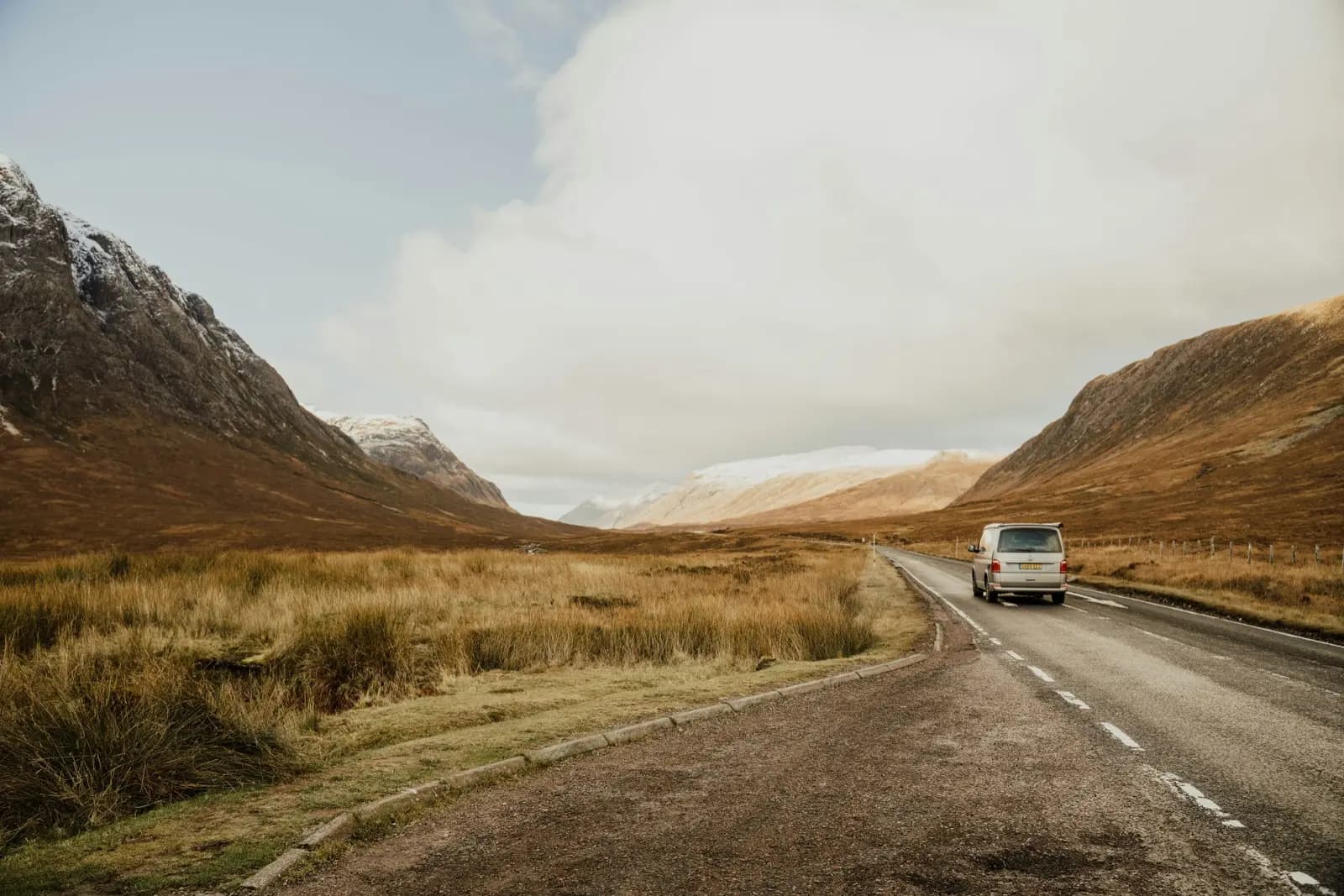 Silver VW campervan driving alone through Glencoe with snow-capped mountains rising on either side