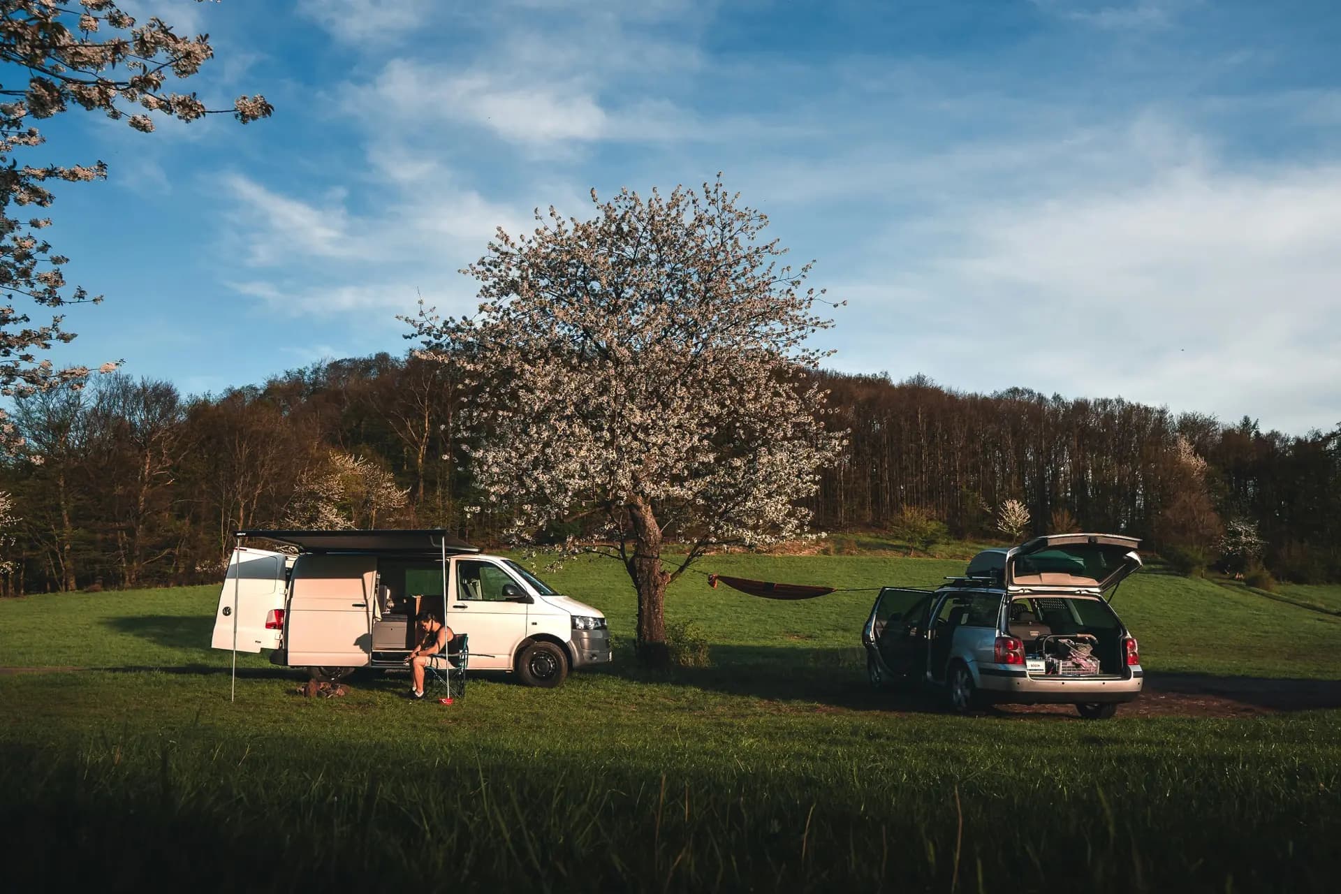 White VW T5 with the awning out in a spring meadow, camping chair set out in the late sun, cherry tree in full blossom behind, a second car parked nearby with a hammock strung between them.