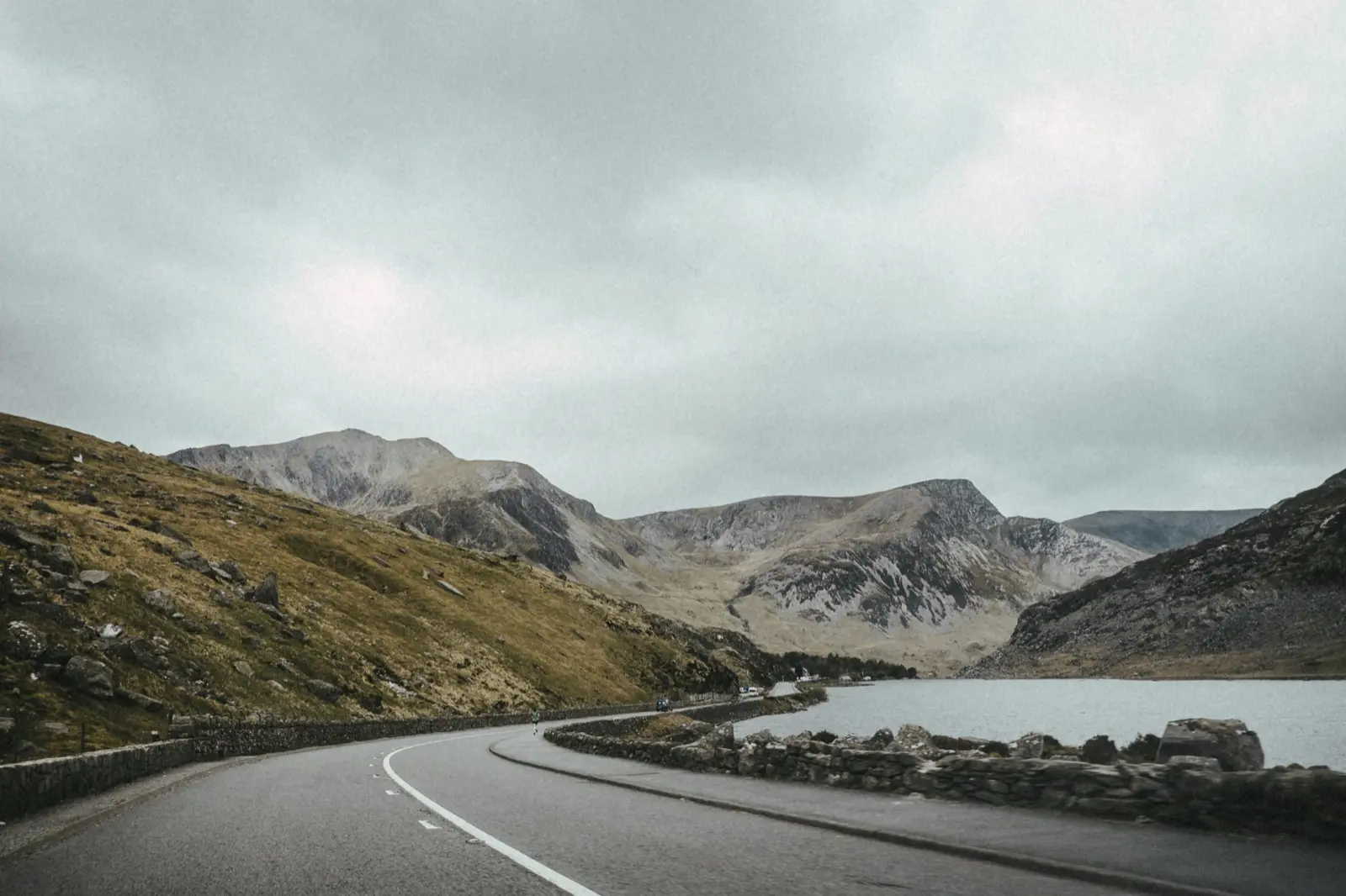 Empty road curving alongside a lake towards rugged grey mountains under an overcast sky in Snowdonia