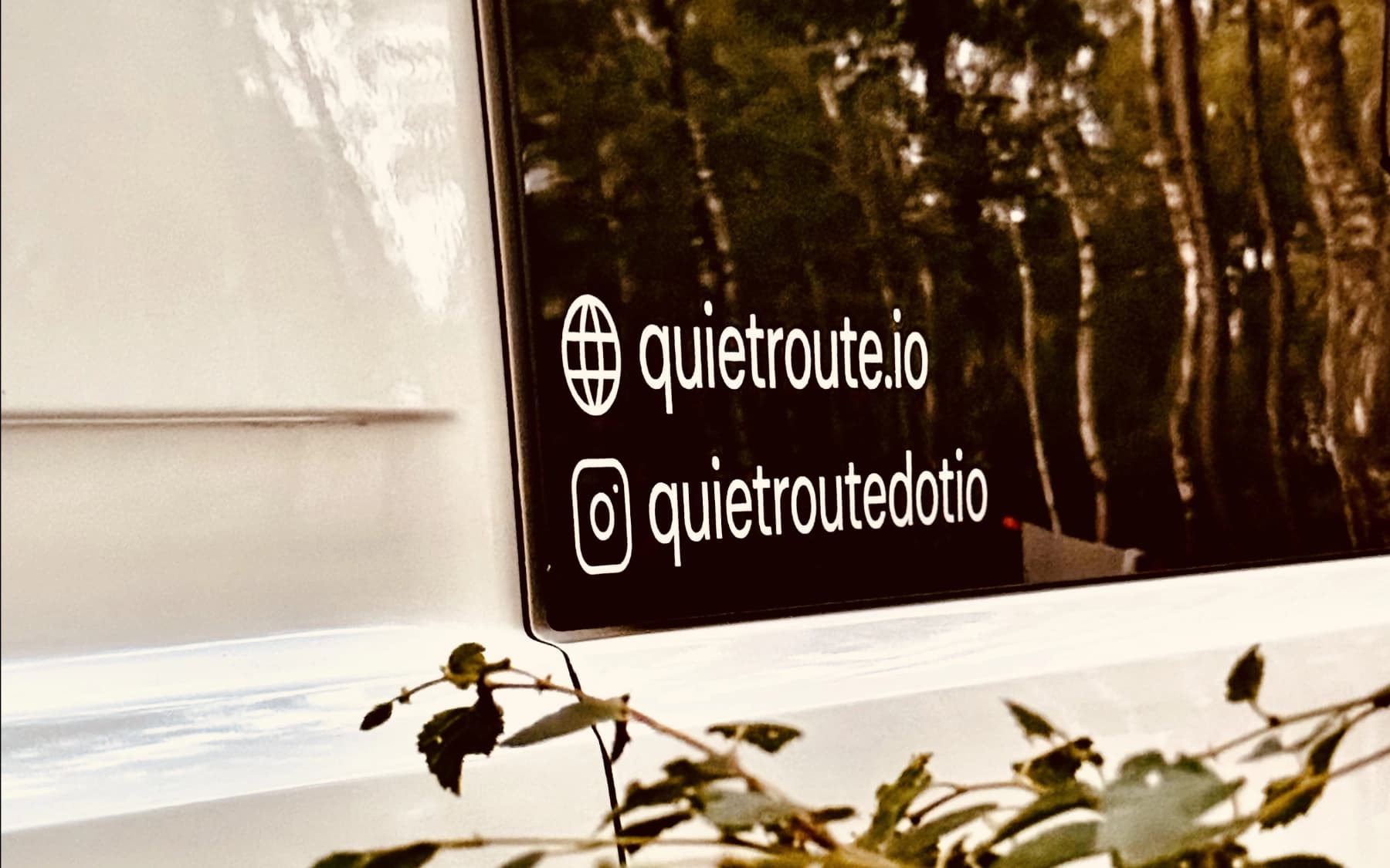 White vinyl decals on a campervan window reading quietroute.io and the Instagram handle quietroutedotio, with woodland reflected in the glass.